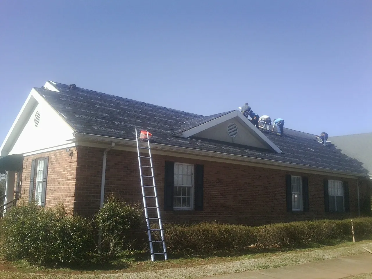 Crew tearing shingles off a brick ranch roof with a ladder set — Metal Roof Repair work by our crew in Calmar