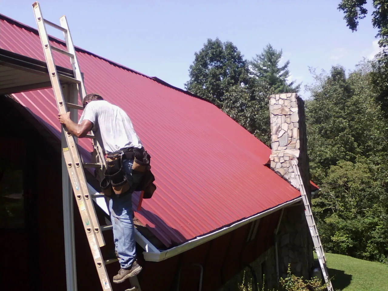 Roofer on a ladder beside a red metal roof and stone chimney — Soffit & Fascia Repair by Roofing Brothers in Calmar